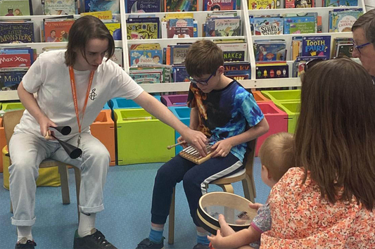 An RSBC staff member in a white top and trousers sits in front of colourful boxes as she helps a brown-haired boy to play the xylophone, with parents and children looking on