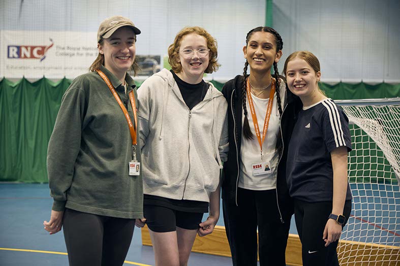 A group of four young women, including two RSBC representatives wearing lanyards, at a residential trip. They're standing in front of goalposts and smiling at the camera happily.