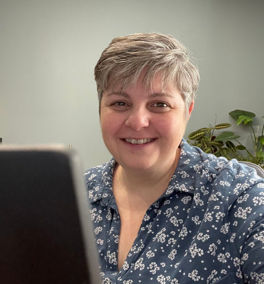 Jeanette, family practitioner, sits in front of a laptop. She is light skinned with short grey hair and wears a blue and white patterned shirt as she smiles broadly at the camera.