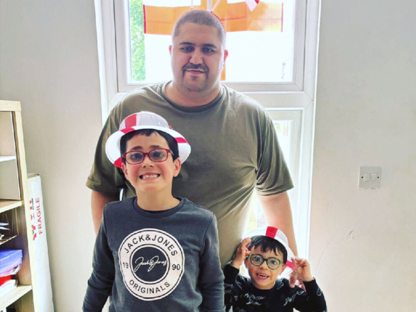 Dad Joe stands behind his sons Noah and Lyle. They are all smiling at the camera, and the boys are wearing England flag hats.