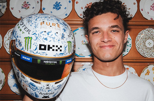 Lando Norris, a young man with curly brown hair and wearing a white T shirt, poses in front of rows of china plates while he holds the special porcelain helmet
