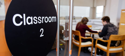 The open door, which says Classroom 2, to a room where a student and teacher sit at a table with their backs toward the camera.