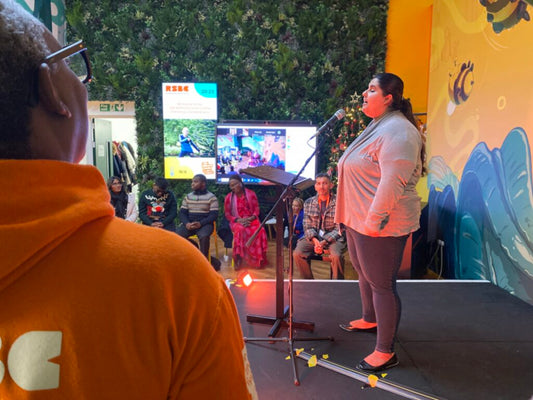 Zenny stands on stage, singing into a microphone at at RSBC's Life Without Limits Centre. An RSBC staff member in an orange top with an RSBC logo and other people watch.