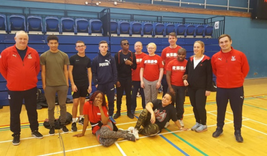 A group of young people and Palace for Life staff in a gym hall stand in a row with two of them sitting on the floor in front, all smiling at the camera