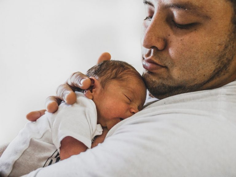 A dark haired, medium skinned man cradles a small child to his chest, looking down at it tenderly.