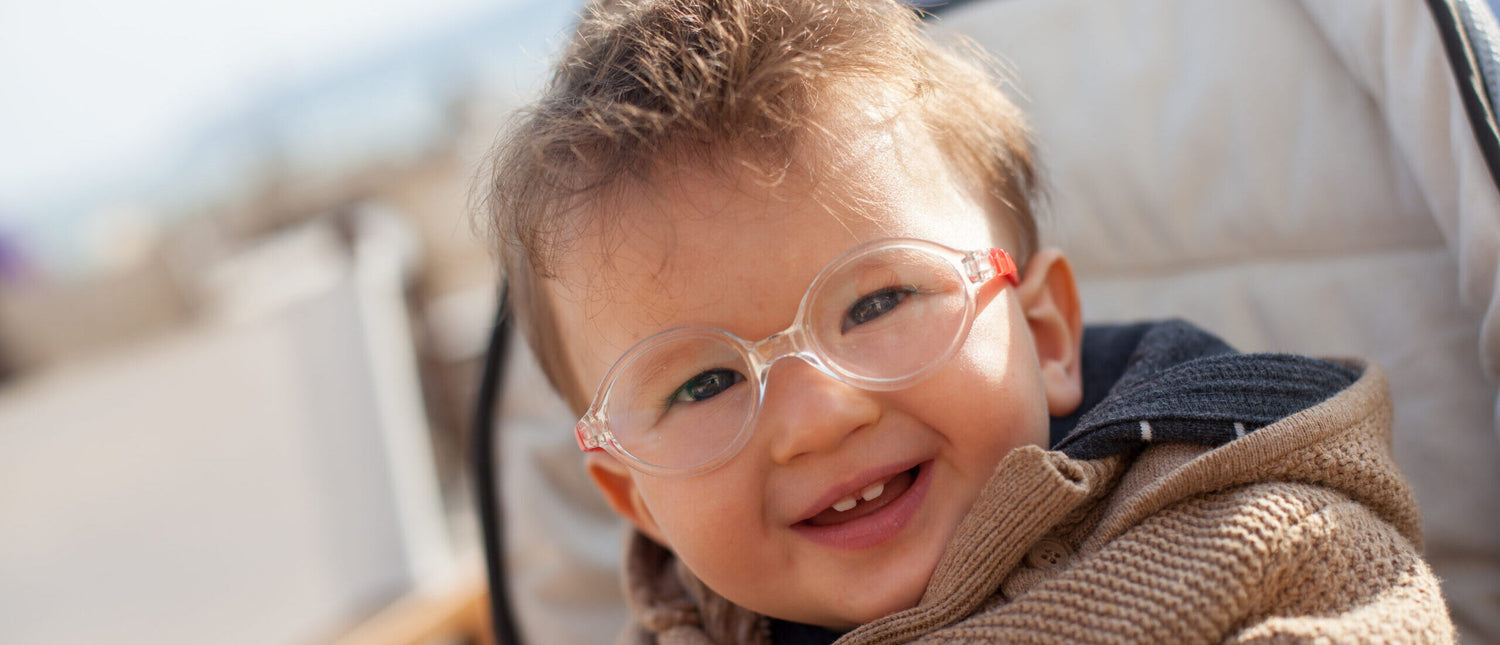 Young boy, around age 4, facing the camera, smiling, wearing circular glasses with an orange frame and a brown knitted textured coat.