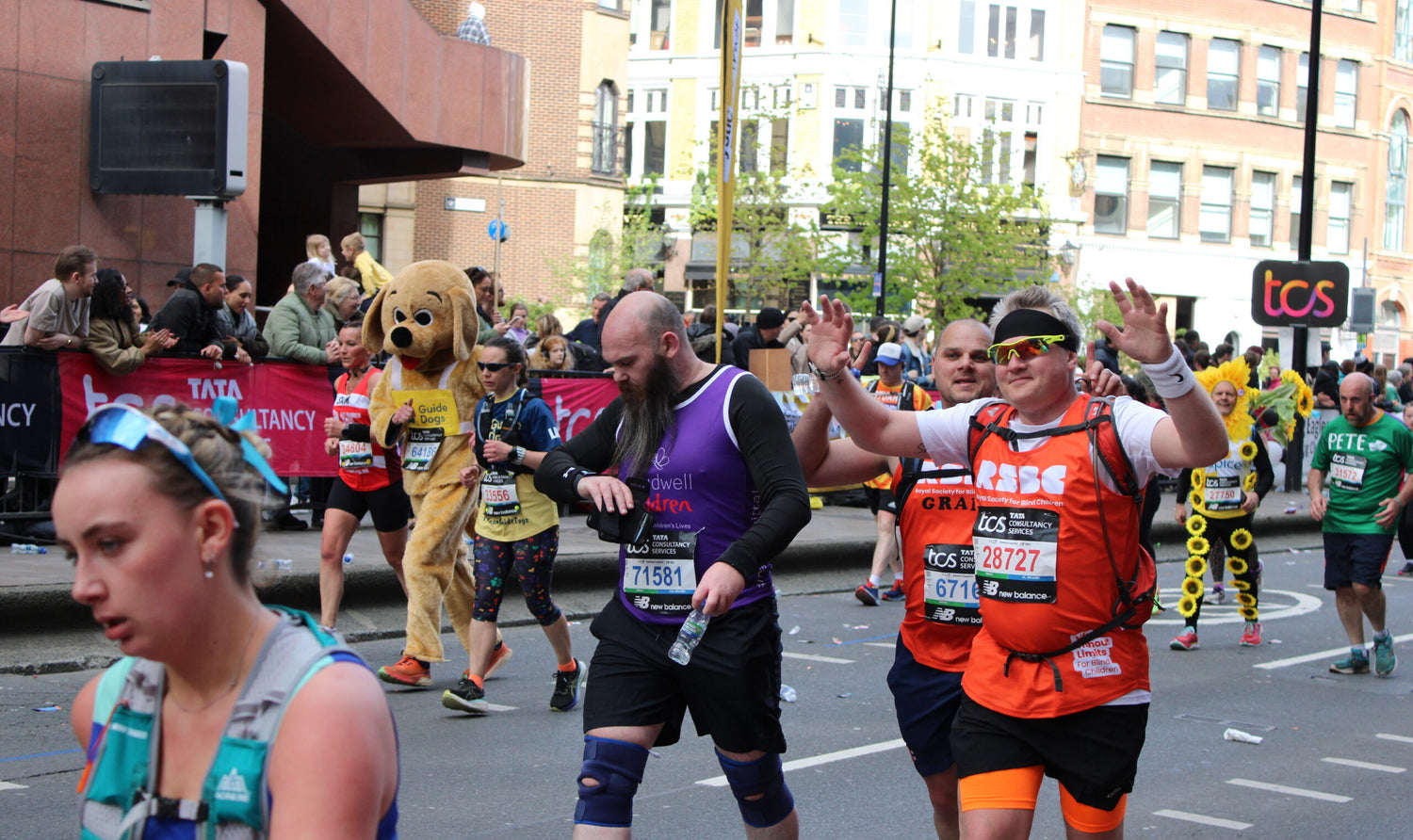 A London Marathon runner in an orange vest and sunglasses has his arms raised in the air celebrating that he is most of the way to the finish line