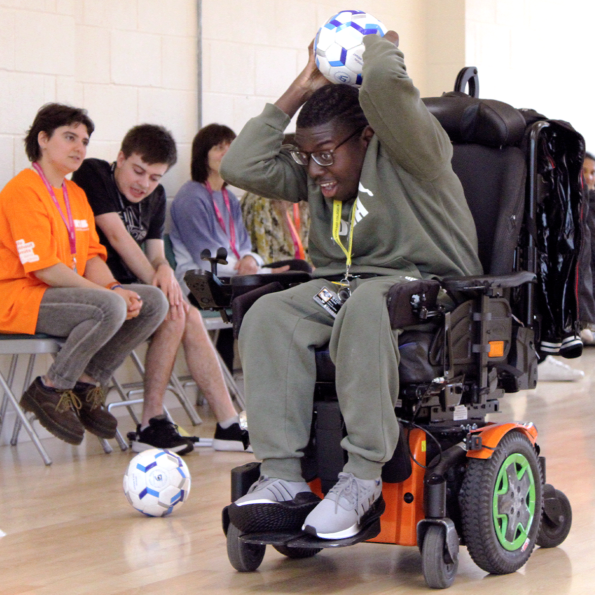 A dark-skinned young boy in a motorised-wheelchair. He is holding a ball above his head, preparing to throw it. His face is full of concentration.