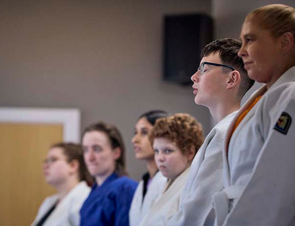A group of young people stood in a line in a Judo session. One young man has his eyes closed, prepararing calmly.
