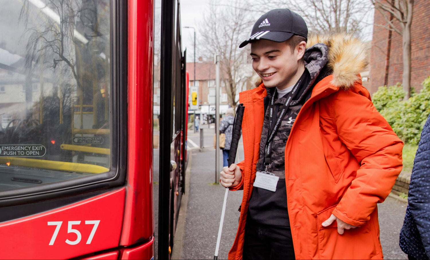 A student wearing an orange jacket and carrying a white cane is about to board a red bus.