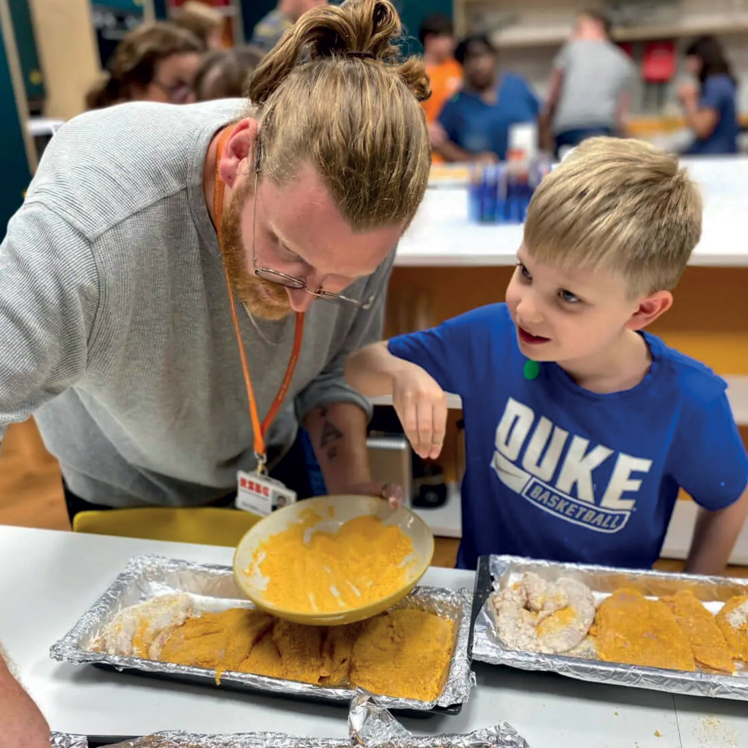 A young boy with blonde hair is having a great time learning to cook in our kitchen, standing next to a male adult who has long hair, a beard and glasses