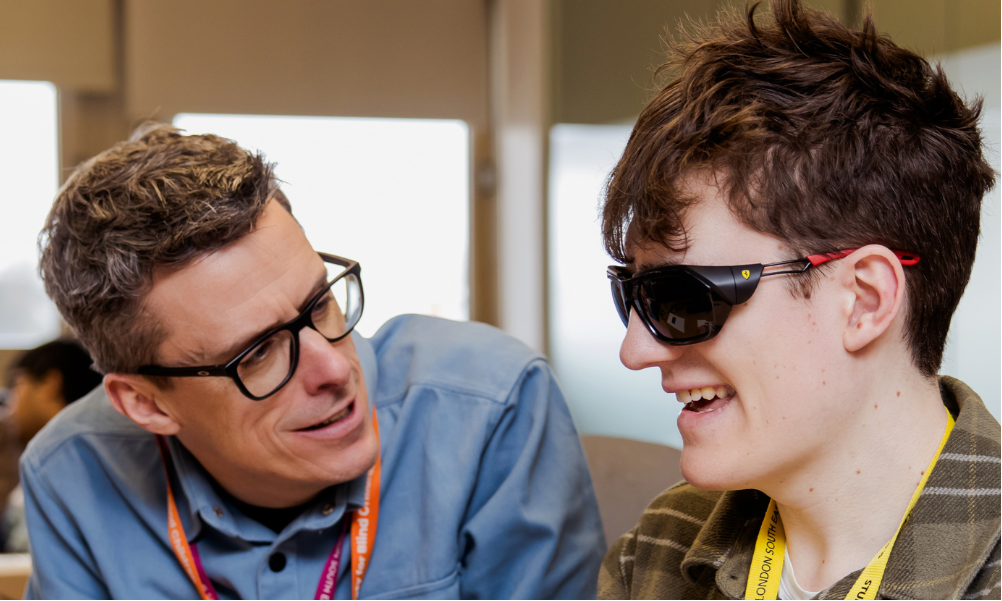 A young man with glasses and his QTVI smiling during a study session