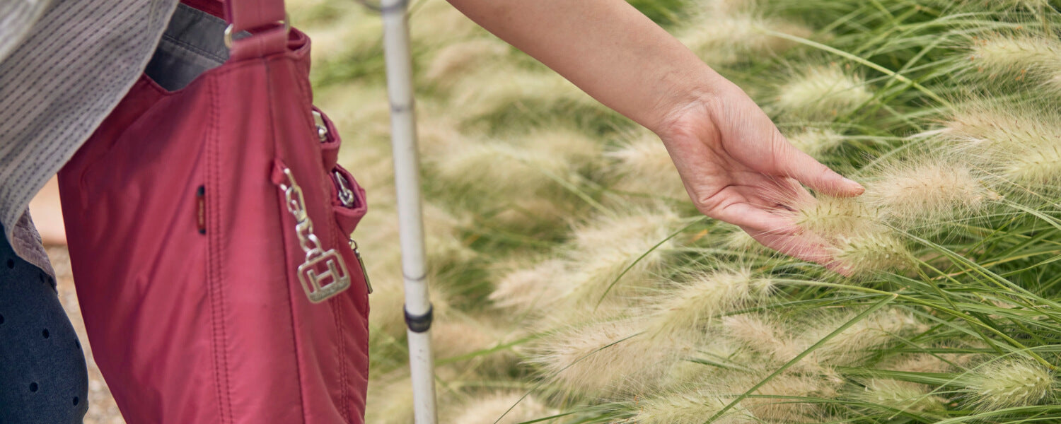 A person with a cane, carrying a pink canvas bag, is touching wheat in a windy field