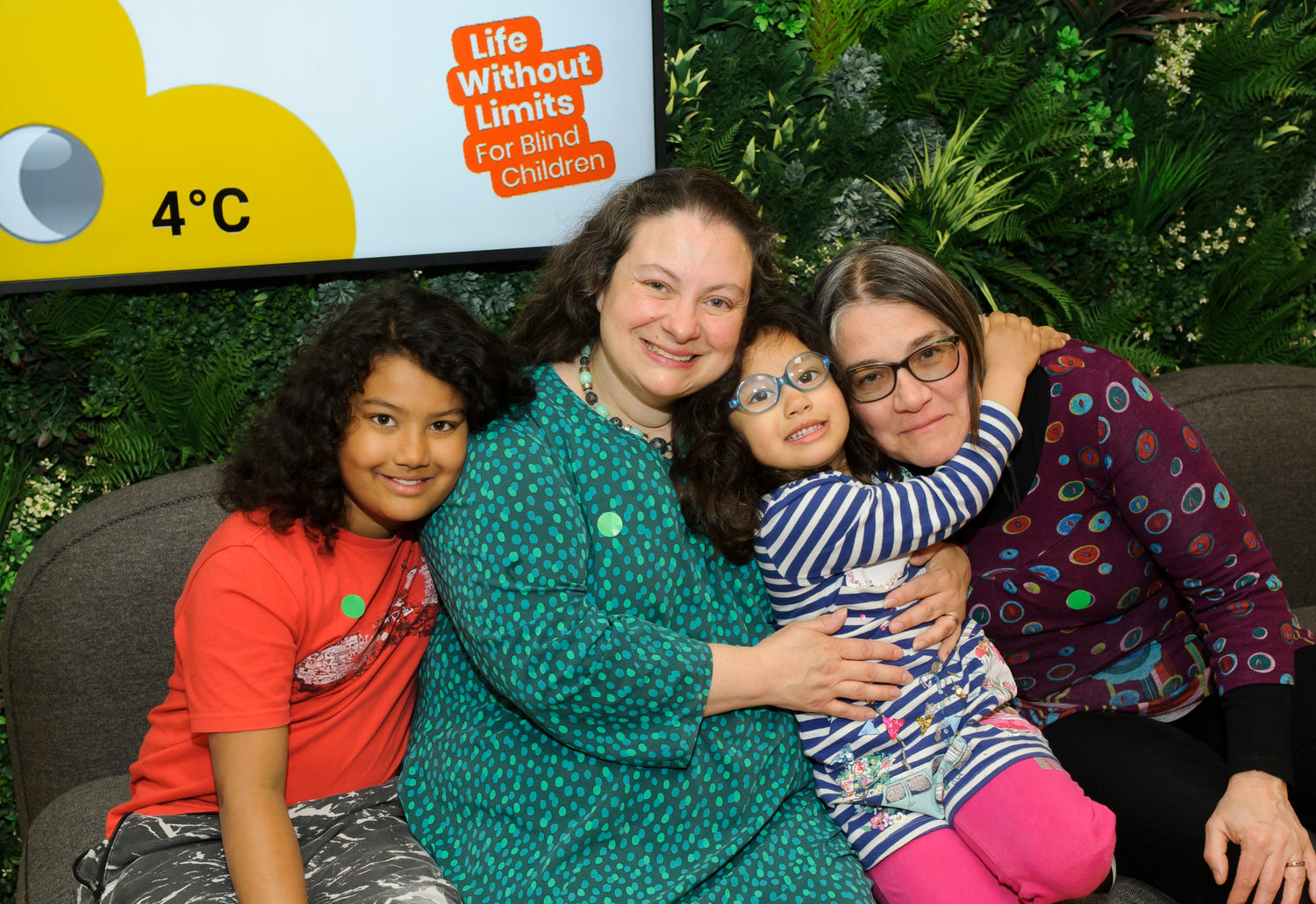 A happy family posing together for a picture. The family, two women with small children either side of them, sit on a sofa in the Life Without Limits Centre with arms around each other smiling brightly.