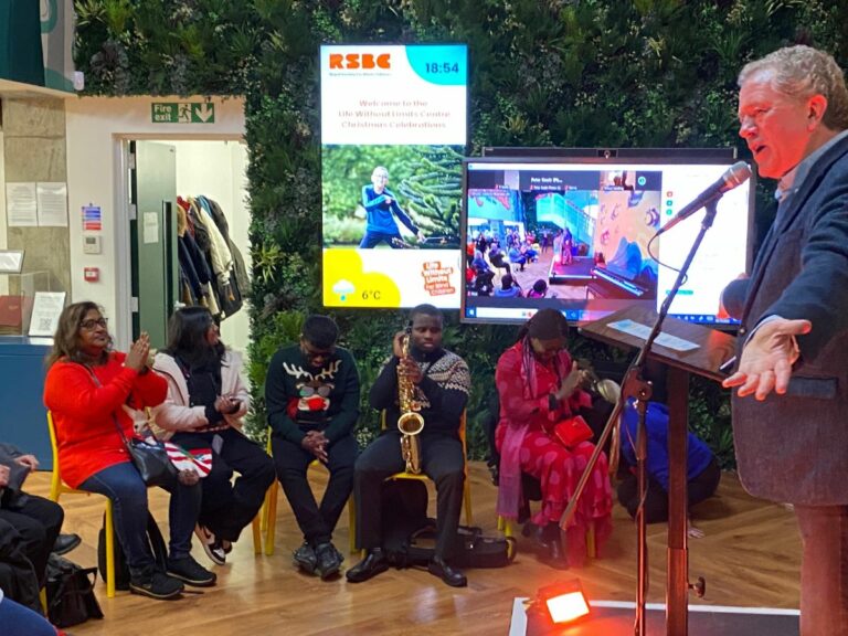 Jon Culshaw stands at a microphone at the Christmas Concert, talking to the attendees while some of the talented young musicians who performed sit and watch him. 