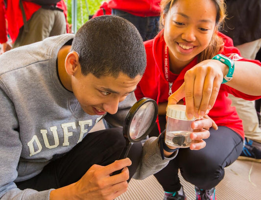 A young dark-haired man in a grey sweatshirt is looking through a magnifying glass at an object in a glass of water, as a volunteer smiles next to him and holds the glass up.