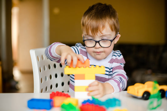 A young boy with auburn hair and black glasses sits on a white chair at a white desk. He's playing with brightly coloured building blocks and a yellow toy car is next to him. His face shows that he's concentrating as he puts the yop block in place.