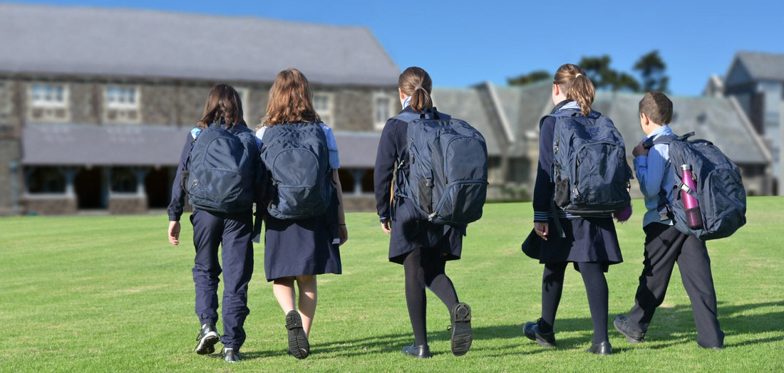 A group of five young secondary school pupils walk across grass to their school, seen from the back