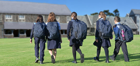A group of five young secondary school pupils walk across grass to their school, seen from the back
