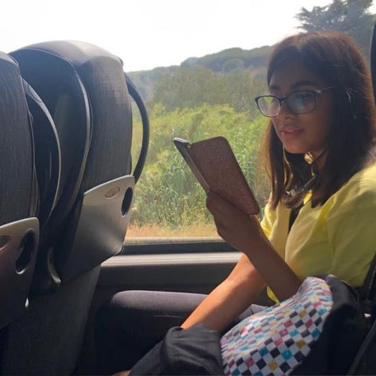 Alyssia, wearing glasses reading a book outdoors with green hills in the background on a sunny day.