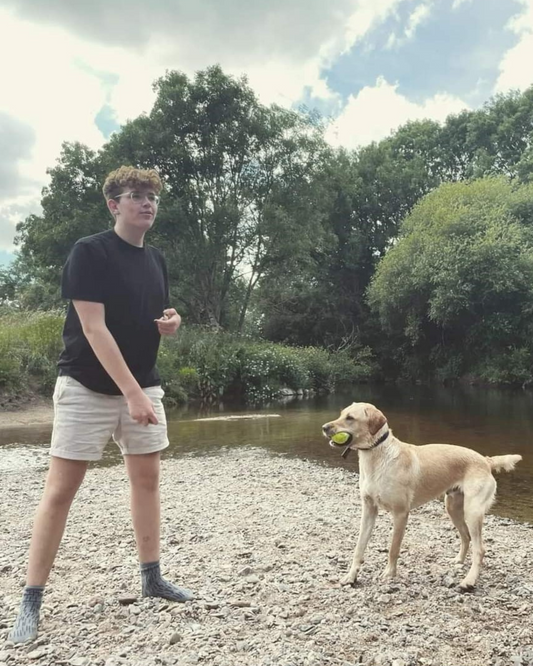 Harvey, a young man with brown hair, wears a black T shirt and biege shorts as he plays ball with Alfie, a yellow labrador, on a sandy riverside.