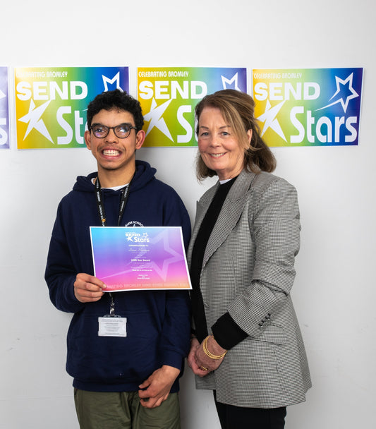 Isaac, a young man with brown hair and glasses, smiles braodly as he stands next to a woman who is also smiling at the camera. Behind them are three posters each coloured yellow, green and blue which say 'SEND Stars' and have a star symbol on them.