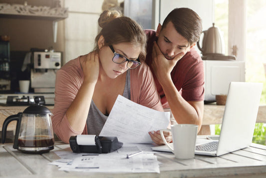 A woman and her male partner are in their kitchen, looking worriedly at a bill with a calculator and a laptop in front of them.