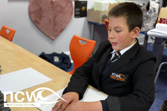 Ed, a New College Worcester student, wearing a dark school uniform, sits at a desk with bright orange chairs in the background, He is concentrating with his fingers on a white sheet of paper as he reds a story to record for the Bedtime Donations app.