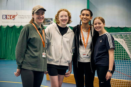 A group of four young women, including two RSBC representatives wearing lanyards, at a residential trip. They're standing in front of goalposts and smiling at the camera happily.