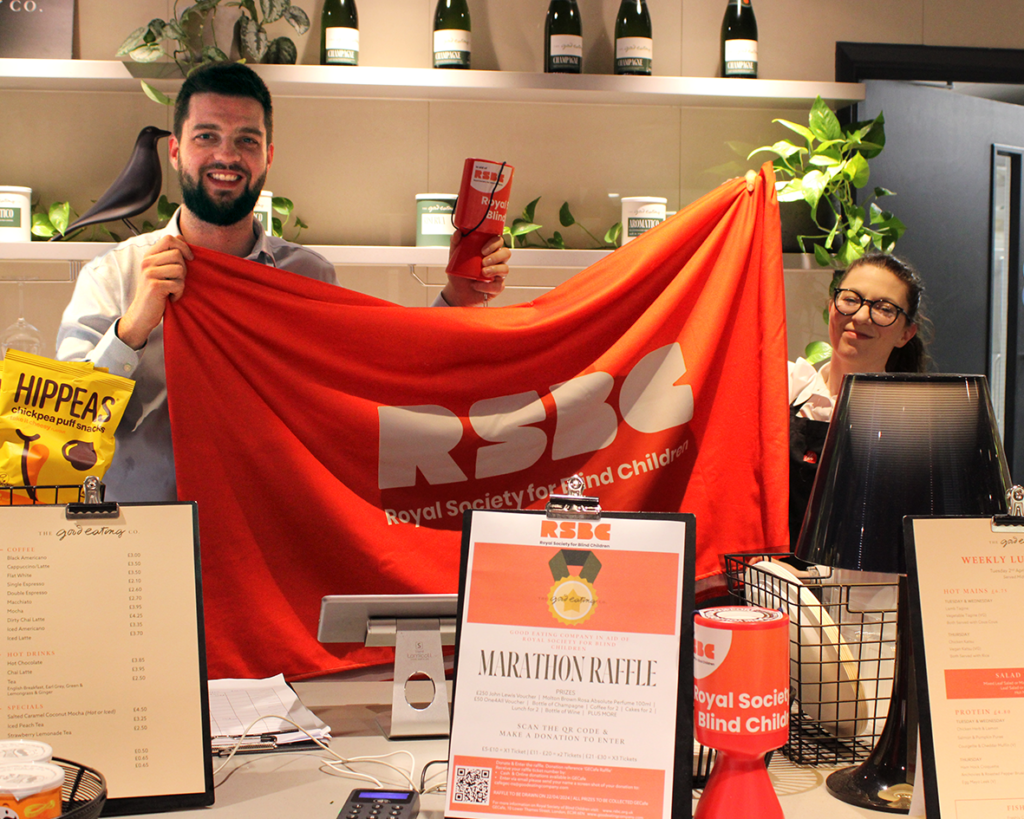 Two members of staff at the Good Eating Cafe stand behind the counter smiling as they hold up an RSBC red banner. In front of them is a sign about the raffle and an RSBC collection tin.