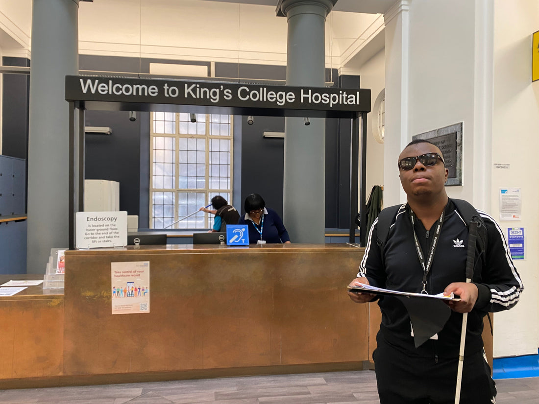  Edesiri, a young man wearing an Adidas sweatshirt and black trousers while holding a clipboard and white cane stands in front of King's College Hospitals reception area with staff behind the desk.