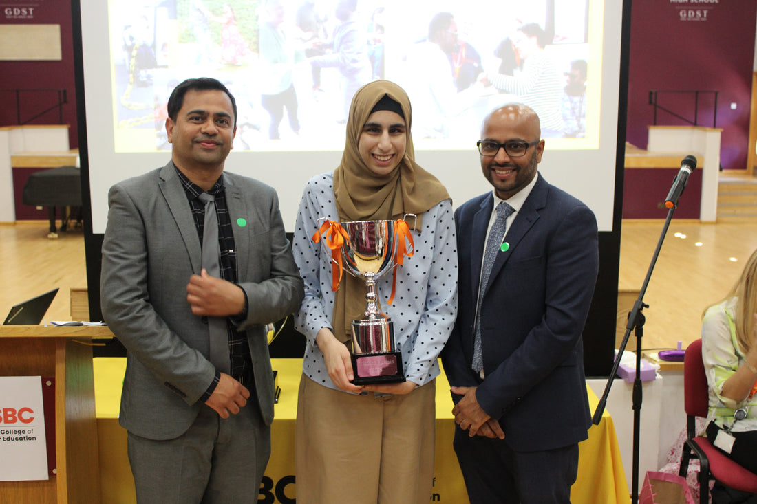 A smiling student holds a big silver cup with ribbons on the handles, and stands between two men in suits. They're all smiling at the camera.