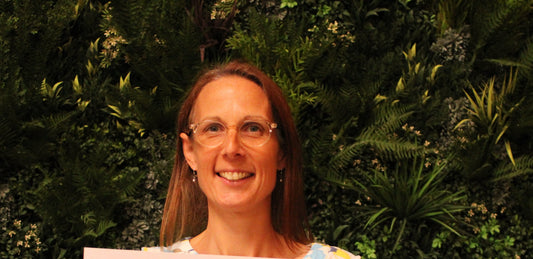 A smiling woman with glasses stands in front of a green foliage wall.