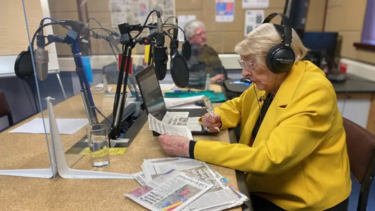 Joy, an older lady sat at a recording booth, is wearing headphones and a bright yellow jacket. She is talking into a microphone reading from printed text.