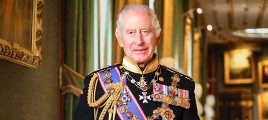 King Charles III, a man with grey hair, stands in an ornate room with many paintings on the walls. He is dressed in royal regalia - a black uniform with gold braid, a purple sash and many colourful medals.