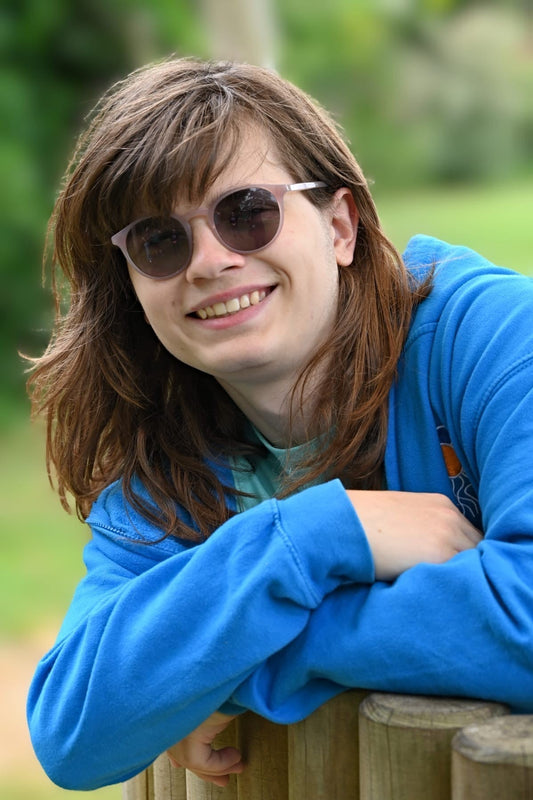 A young man with long brown hair wears sunglasses and a blue hoodie. His arms are crossed as he leans on a fence and smiles broadly at the camera.