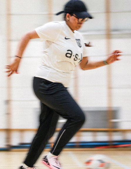 A blurred action shot of a young woman running inside a gym hall. She wears a black skip cap, white T shirt, black jogging trousers and white trainers and looks determined.