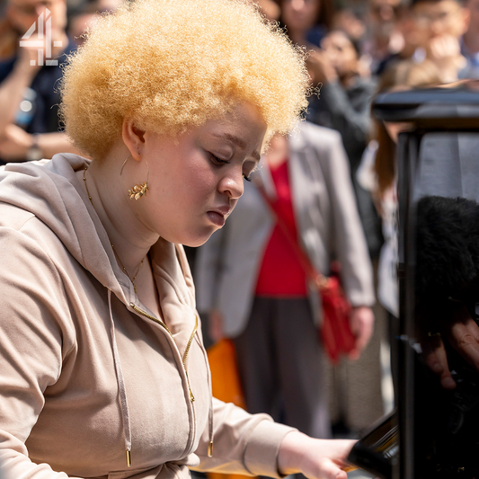 A photo of Precious, playing the piano on Channel 4's The Piano. She has a blonde tight-curled afro and is wearing a light brown hoodie as well as gold hoop earrings. There is an audience standing to the left of her watching Precious play.