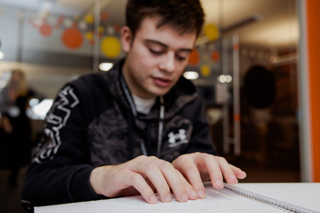  A young man wearing a black and camouflage-print hoodie reads Braille in a bright classroom.