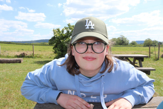 Raffy, A light-skinned young boy wearing a blue hoodie, khaki cap, black framed glasses and black watch. He is leaning on a picnic bench with fields in the background.
