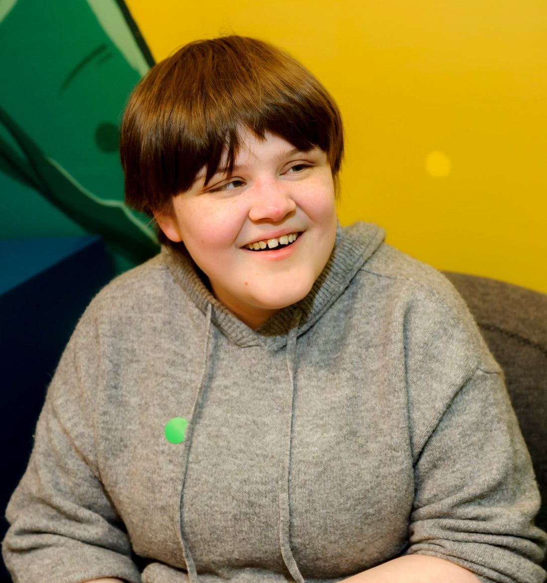 Rona, wearing a grey hoodie, smiling brightly, sitting on a grey couch in a room with vibrant yellow walls.
