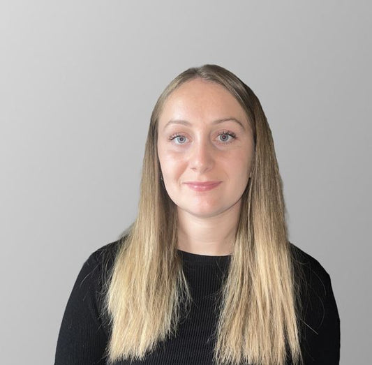 Rosie, a woman with long blonde hair in a middle parting, wearing a black long sleeved top, looks into the camera with a neutral expression.