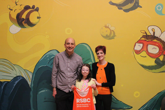 Two adult, Michael and Sarah a child, Rita holding an RSBC running vest in front of a colorful bee-themed wall.
