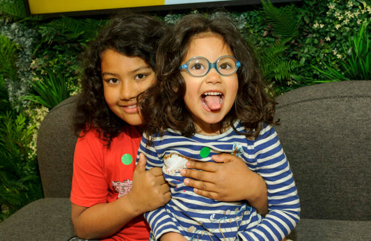 Two young girls with shoulder length dark hair sit on a grey sofa. The older girl has her arms around the younger one, who has blue glasses on and sticks her tongue out at the camera.