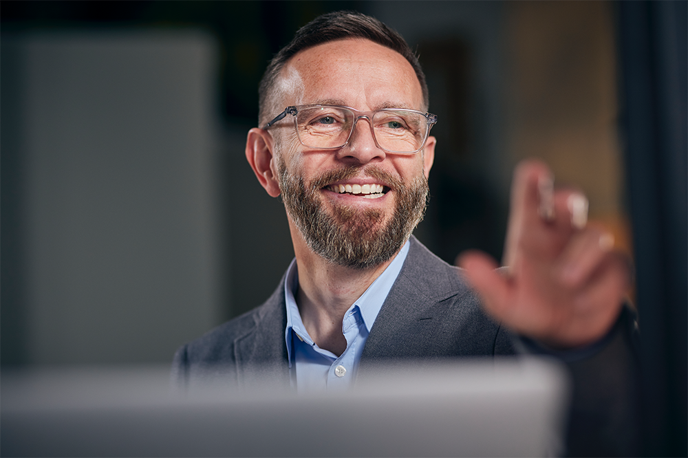 A man with a beard wears glasses, an open necked blue shirt and a grey jacket. He's looking to the right of the image, smiling, with his hand outstretched as if talking animatedley.