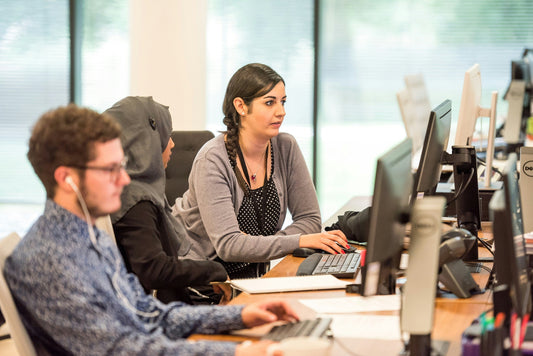 Three people sit in front of computer monitors in an office environment.