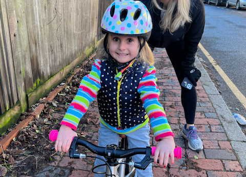 Beatrice, a blonde haired young girl, is on her bike. She wears a light blue helmet with coloured dots, a multi-coloured stripey stop and a blue and white spotty gilet and grins widely, with her hands on the handlebars.