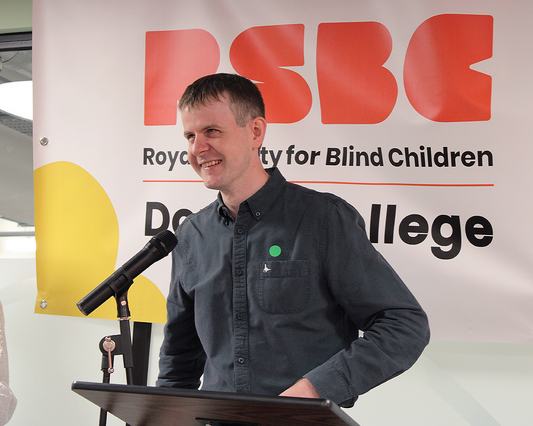 Callum, a light skinned man with dark hair, wearing a grey long sleeved shirt with black buttons. He is standing behind a podium lecture desk, 