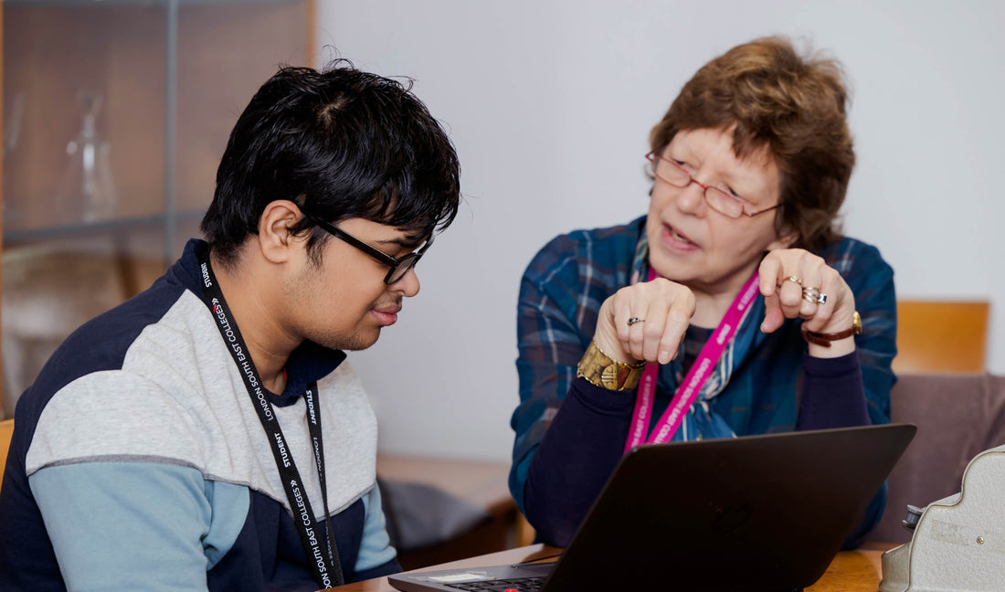 A Dorton College student with black hair and glasses sits at a table next to an RSBC staff member. She is wearing glasses and a blue checked top, and is explaining something to him as they sit in front of a laptop.