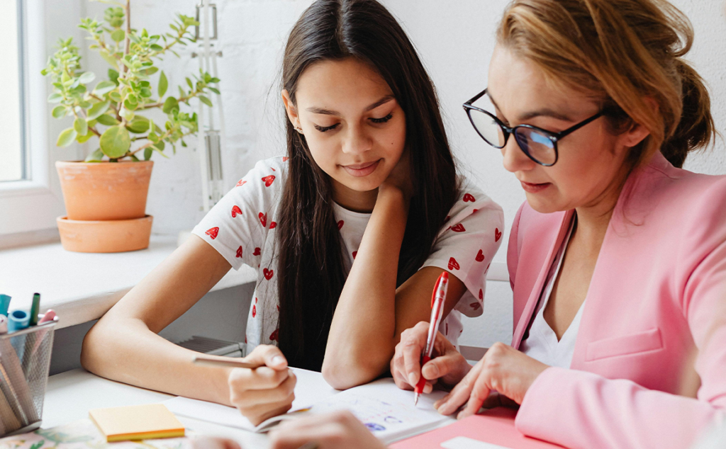 A young woman with long dark hair sits beside a woman with glasses and a pink jacket at a table. They both hold pens, and are working on schoolwork